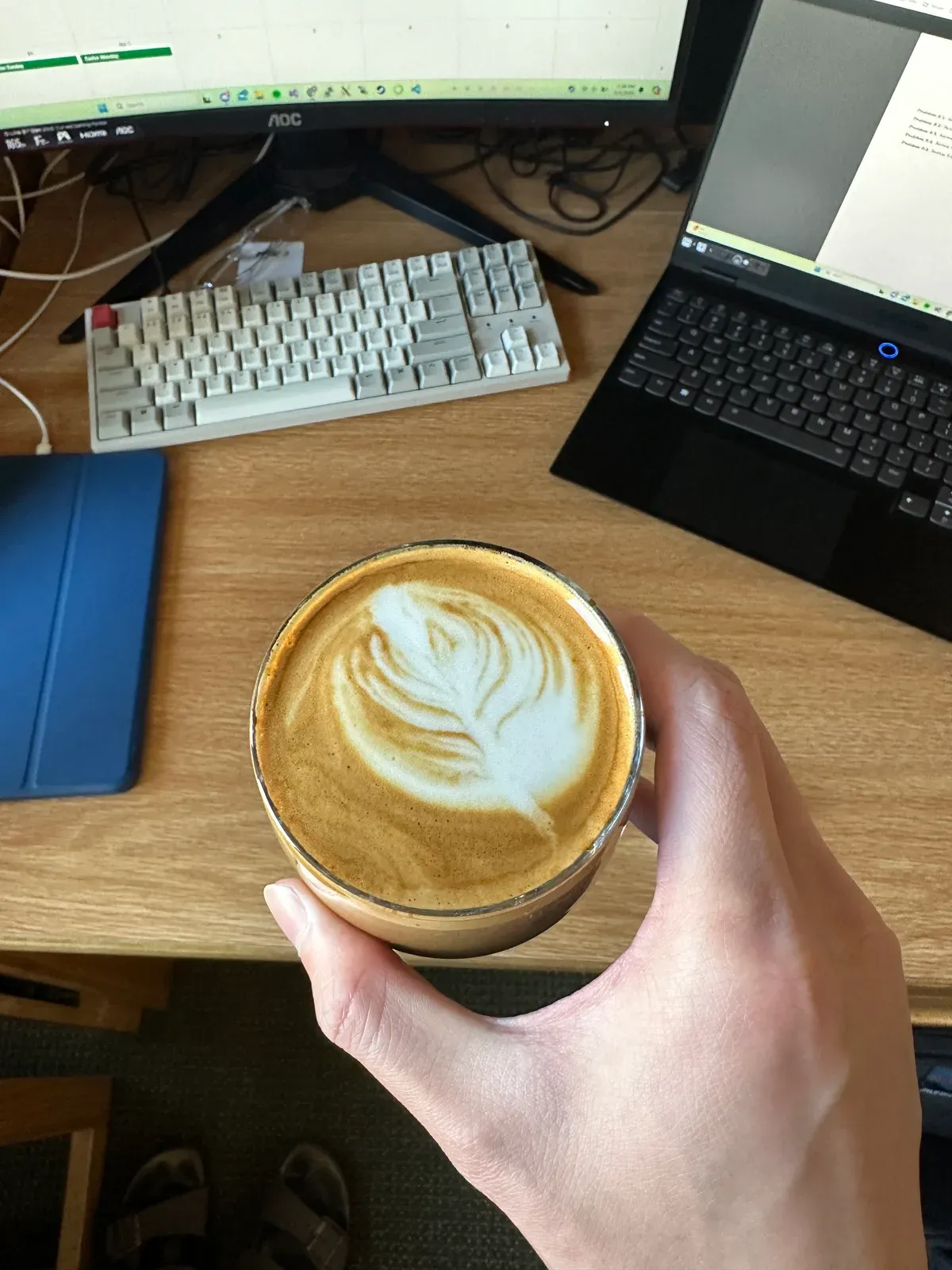A latte with a rosetta poured into a glass, held over a desk with a mechanical keyboard, monitors, and a laptop